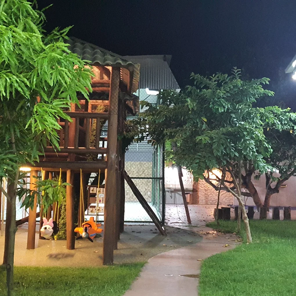 Playground de madeira em formato de casa na árvore, com balanços, iluminado à noite no jardim da Hari Raya Eventos