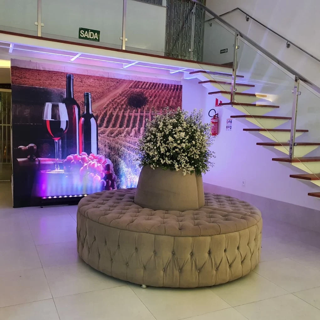 Hall de entrada com puff redondo capitonê, vaso de flores brancas, escada de vidro e madeira e painel iluminado