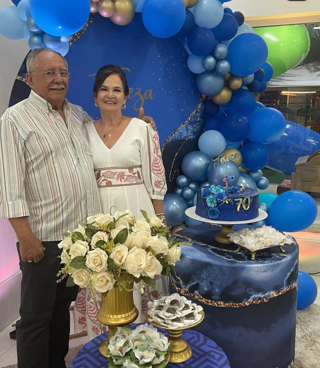 Casal sorrindo em frente a uma decoração azul e dourada com balões e um bolo com o número 70