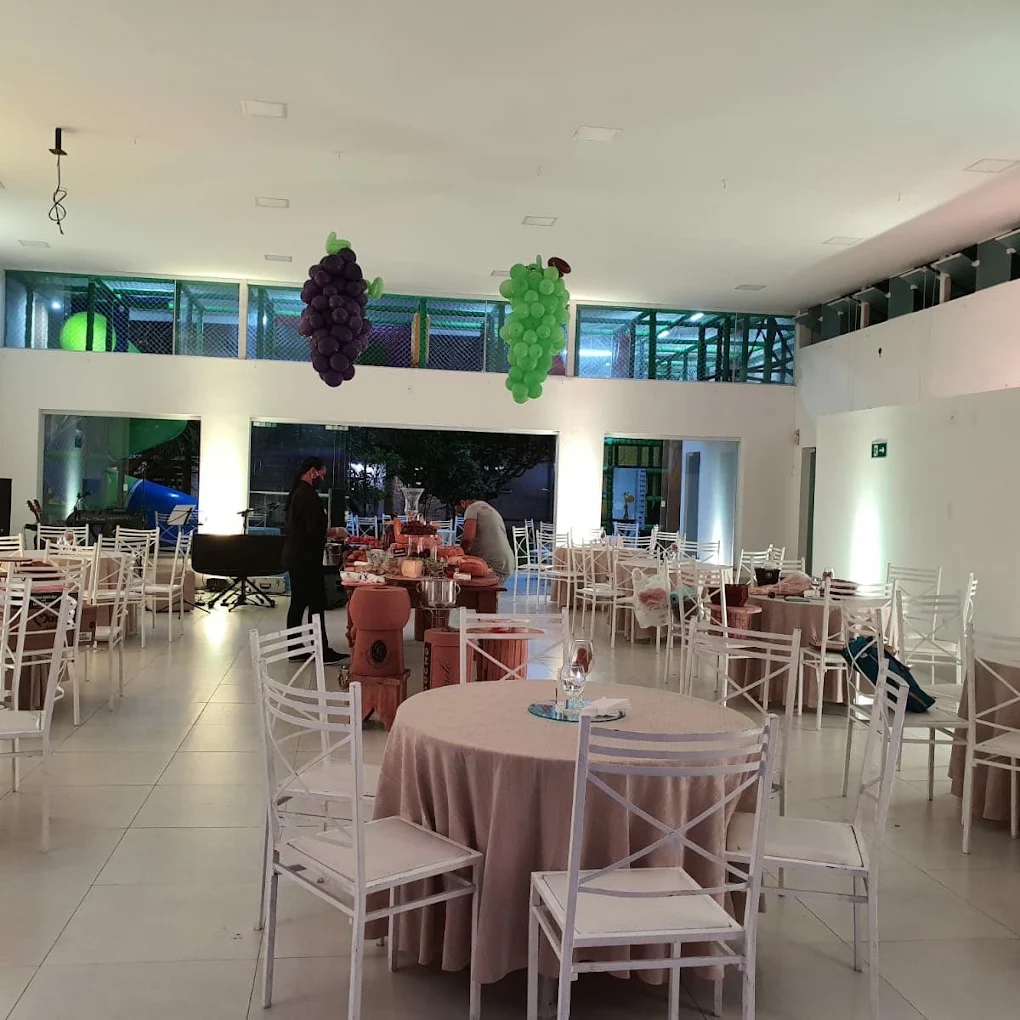 Salão interno montado com mesas redondas, topos de balões e área de buffet, com o playground visível no mezanino