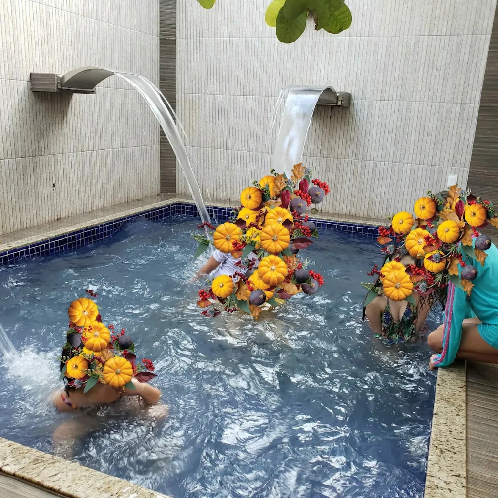 Crianças brincando na piscina sob duas cascatas de água, com arranjos de flores e abóboras na borda