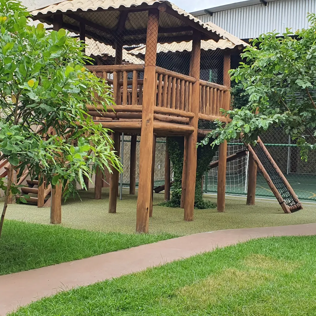 Casa na árvore de madeira com escada e rede, cercada por grama, árvores e caminho de concreto, ao lado da quadra