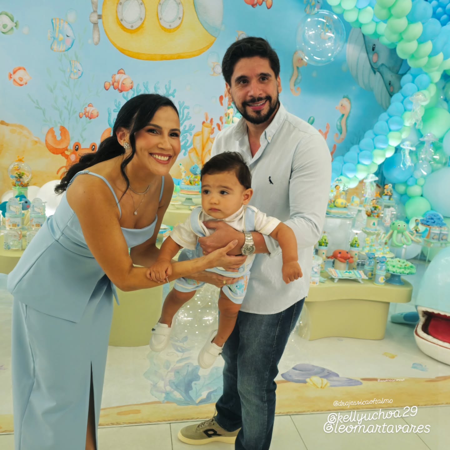 Família em festa com tema fundo do mar, cenário ilustrado e arcos de balões em tons de azul e verde