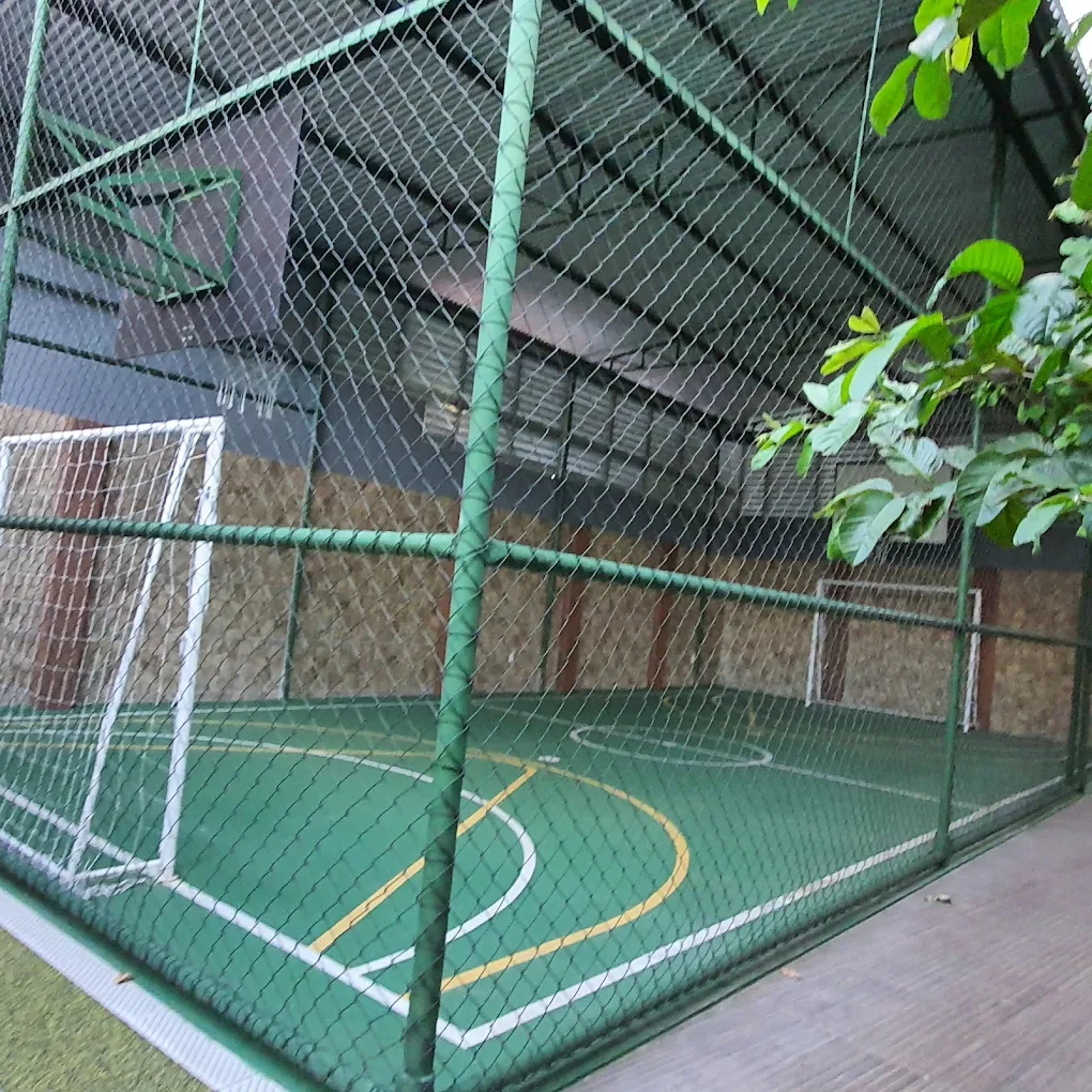 Quadra poliesportiva coberta com piso verde, traves e tabela de basquete, cercada por telha verde