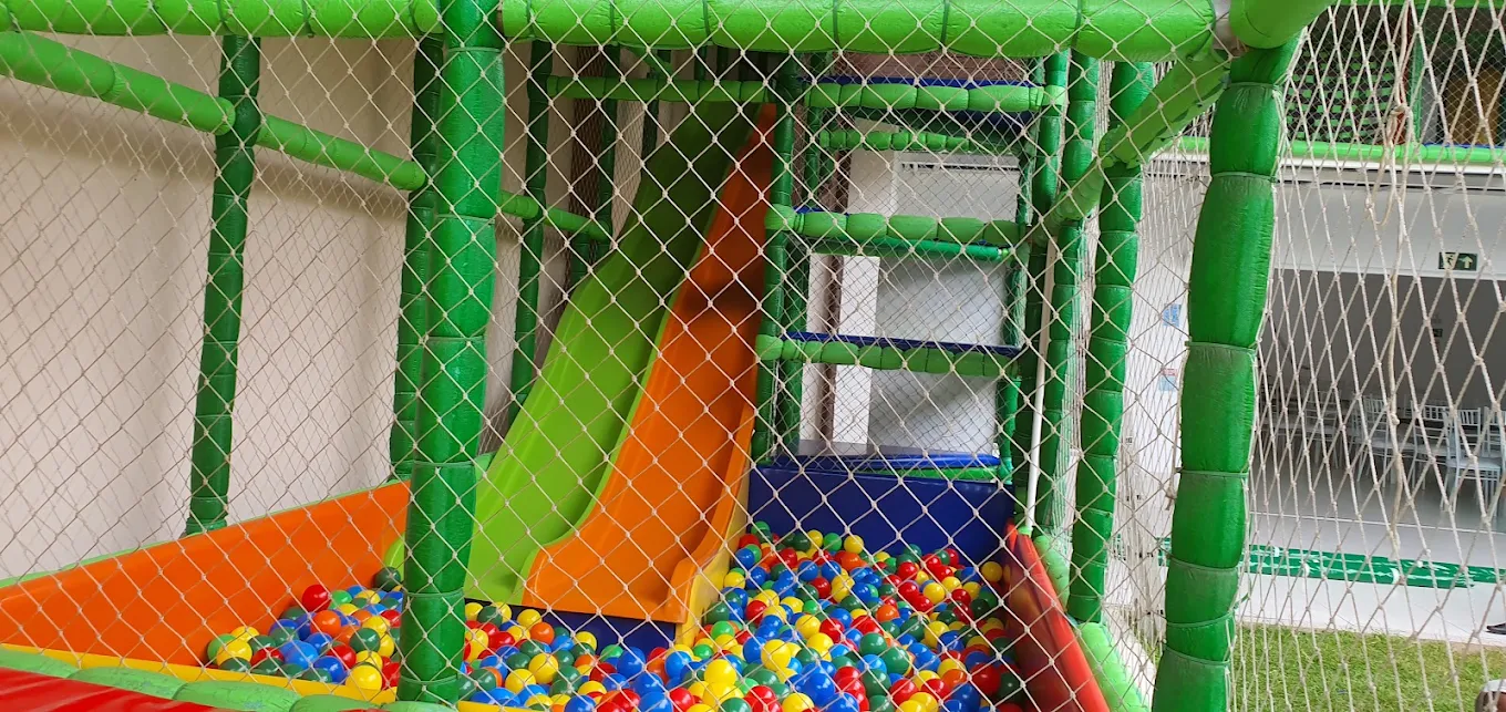 Brinquedão coberto com escorregadores verde e laranja e piscina de bolinhas coloridas, protegido por rede
